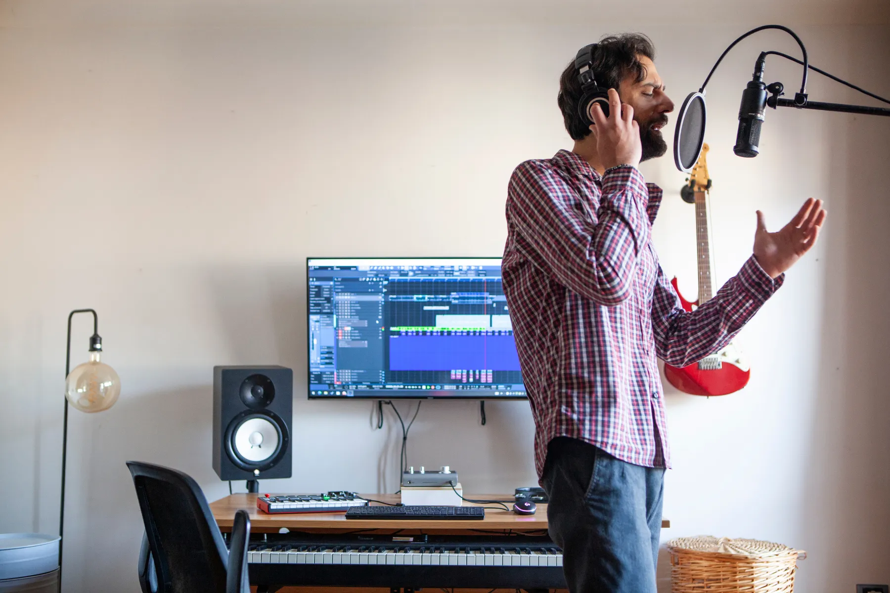 Man recording vocals in a home music studio with microphone, headphones, monitor speakers, and digital audio workstation on screen.